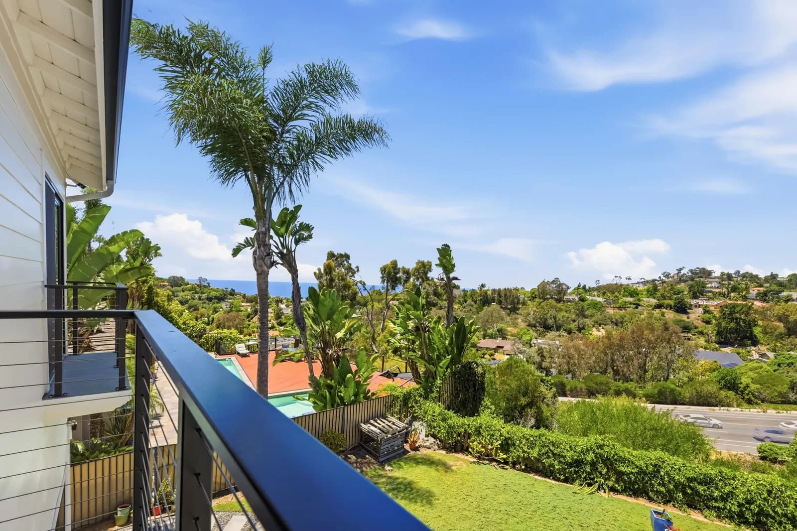 A cable-rail balcony overlooking palm trees, a backyard lawn, rooftops, and the ocean on the horizon.