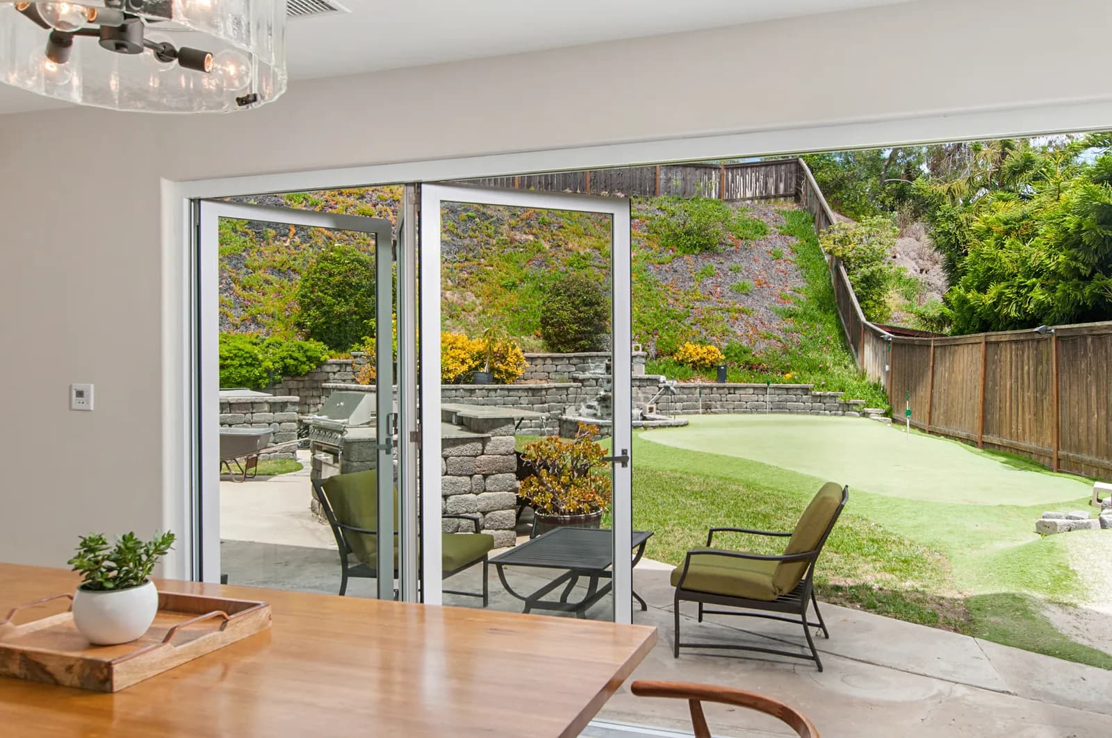 Open glass bifold doors leading from a dining area to a backyard with a putting green, stone retaining walls, and built-in grill.