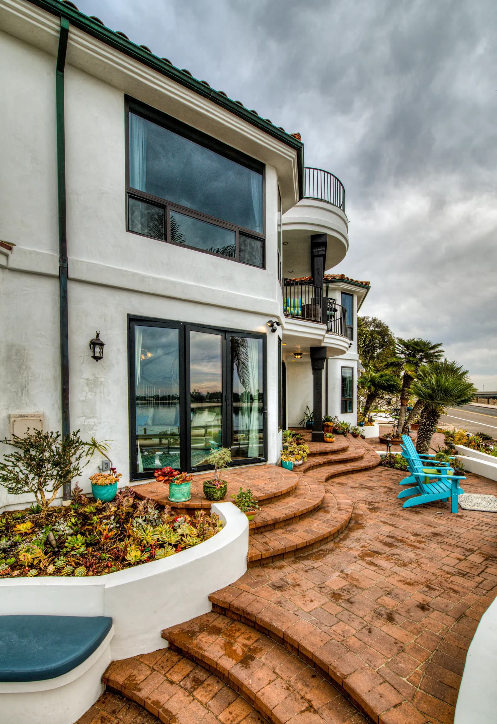 Curved brick paver patio and steps beside a white two-story home with balconies, palm trees, and turquoise chairs.