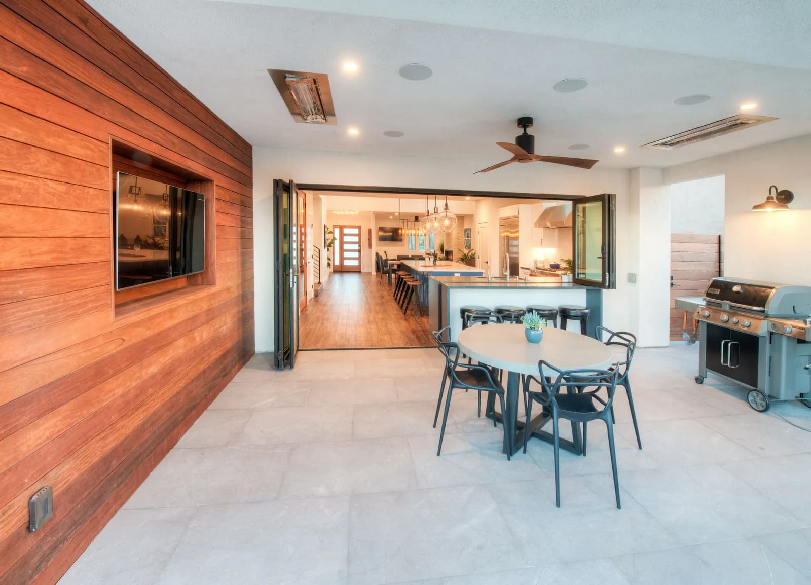 Covered outdoor living room with wood accent wall, TV, grill and folding doors into the kitchen.