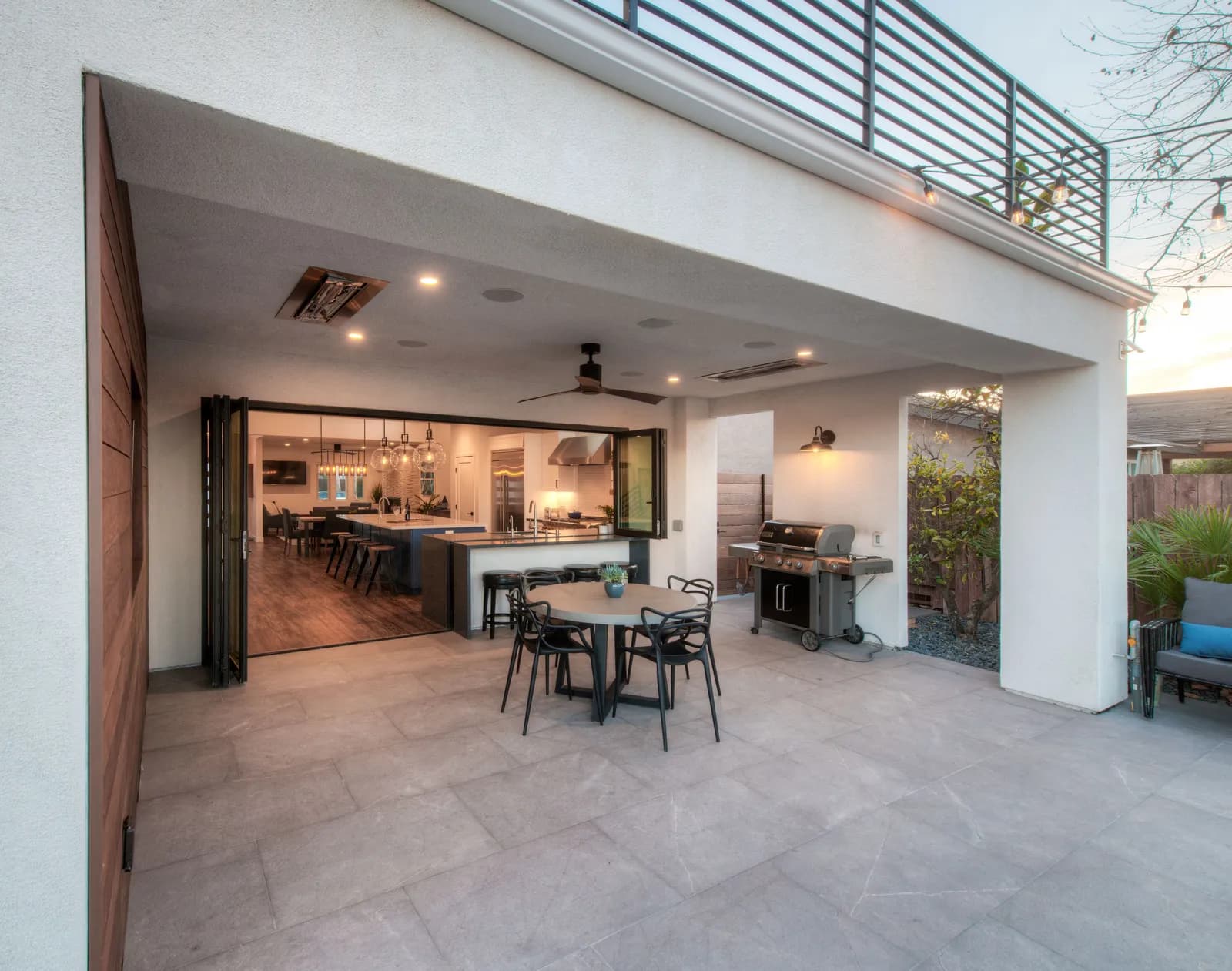 A covered patio with large tile pavers, a round dining table, a grill, string lights, and folding glass doors opening to the kitchen.