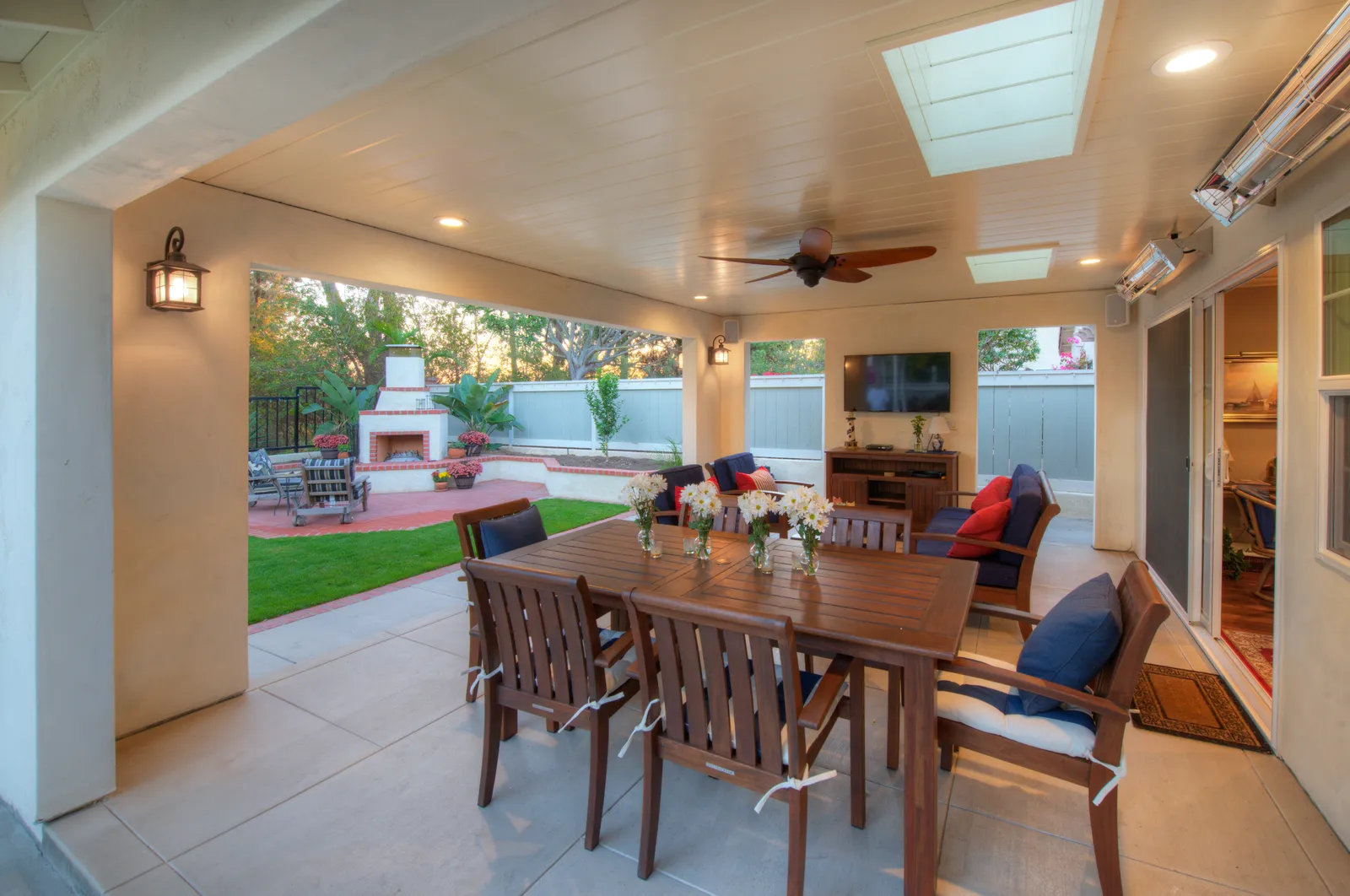 Covered patio at dusk with a wood dining set, ceiling fan, mounted TV, heaters, and an outdoor fireplace beyond.