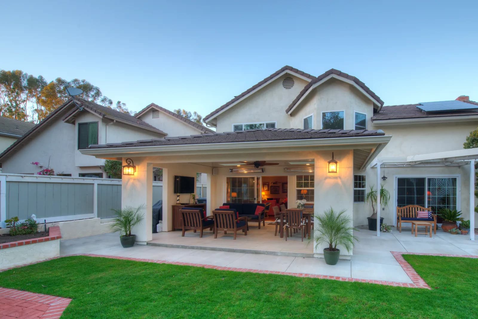 Covered backyard patio at dusk with wood seating, dining area, mounted TV, and manicured lawn.