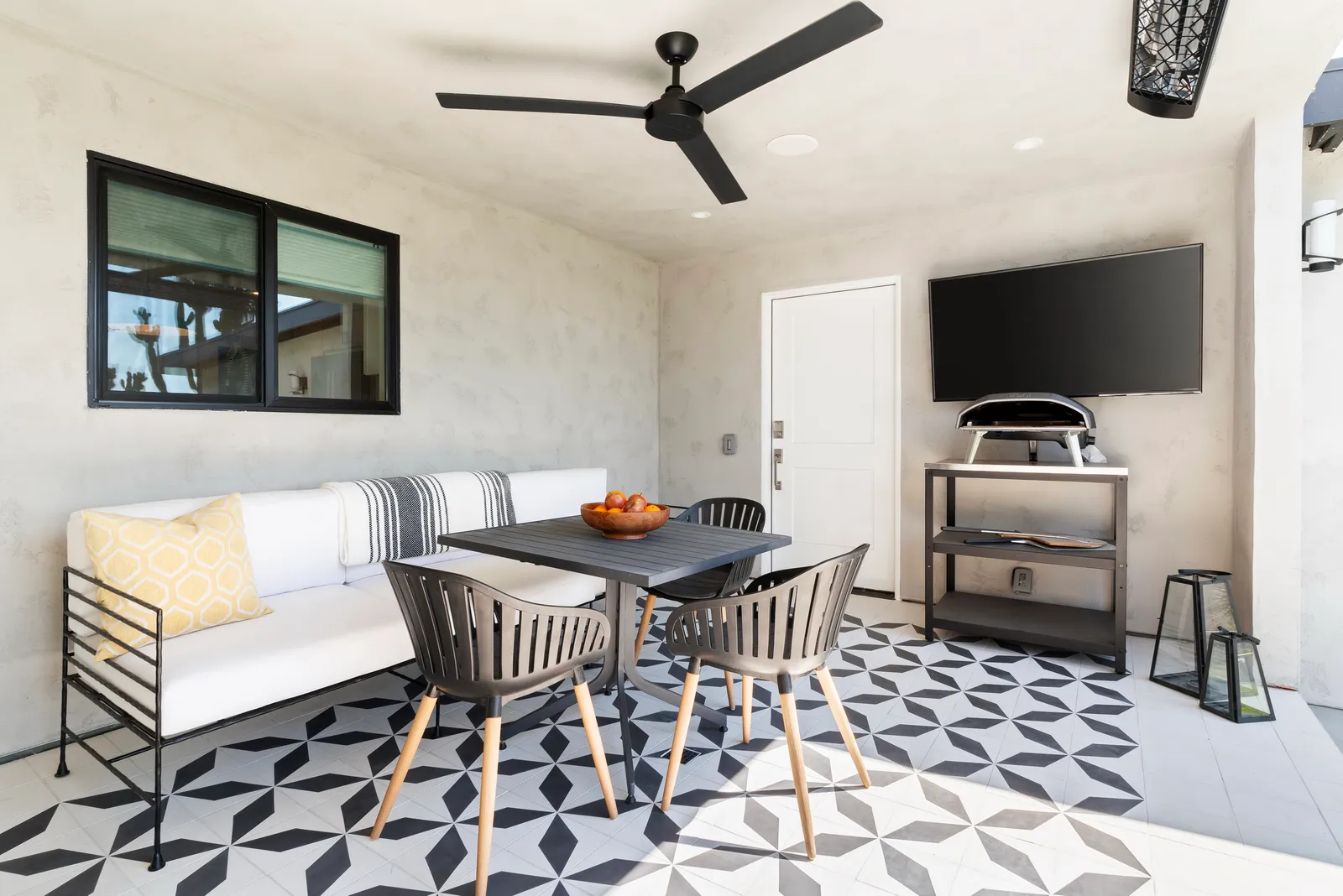 Covered patio with a white outdoor sofa, black dining set, patterned cement tile floor, wall-mounted TV, and pizza oven.