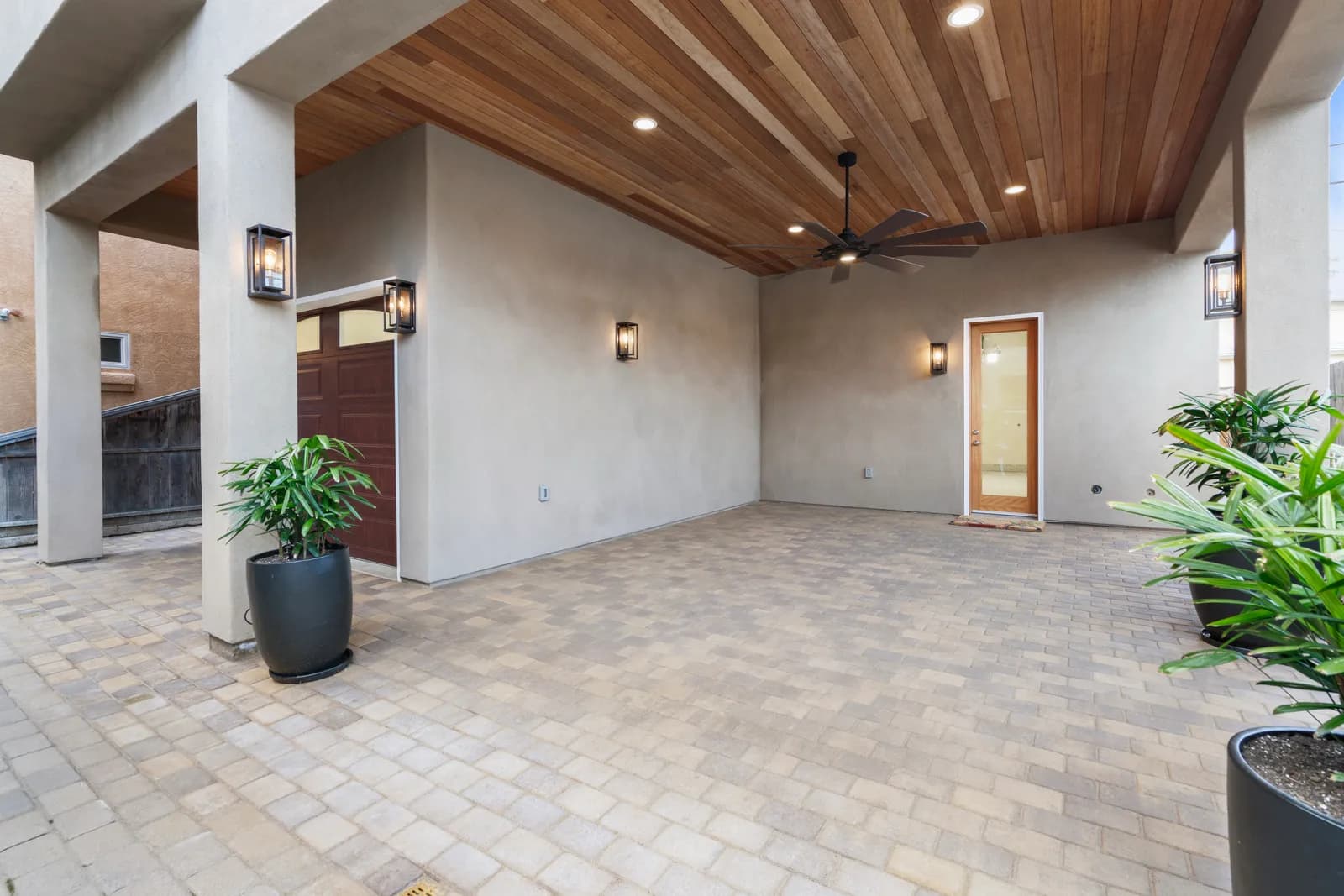 Covered outdoor patio with a stained wood plank ceiling, ceiling fan, lantern sconces, and paver flooring.