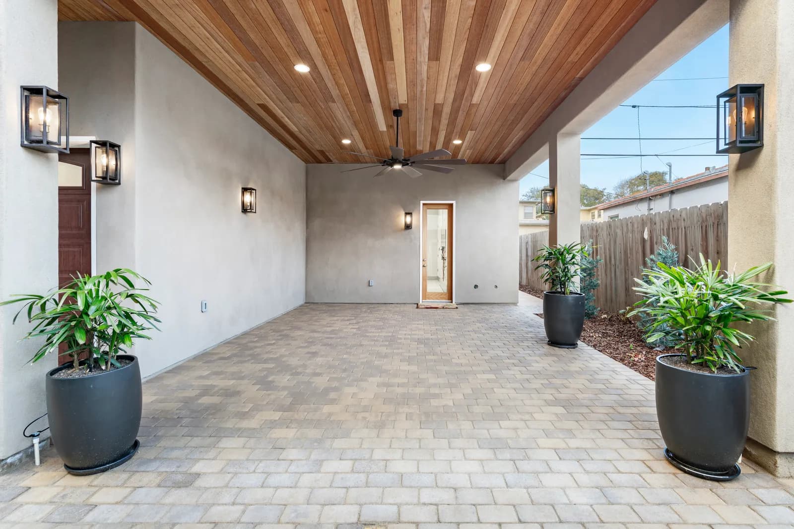 Covered outdoor patio with a tongue-and-groove wood ceiling, ceiling fan, lantern sconces, paver floor, and potted palms.