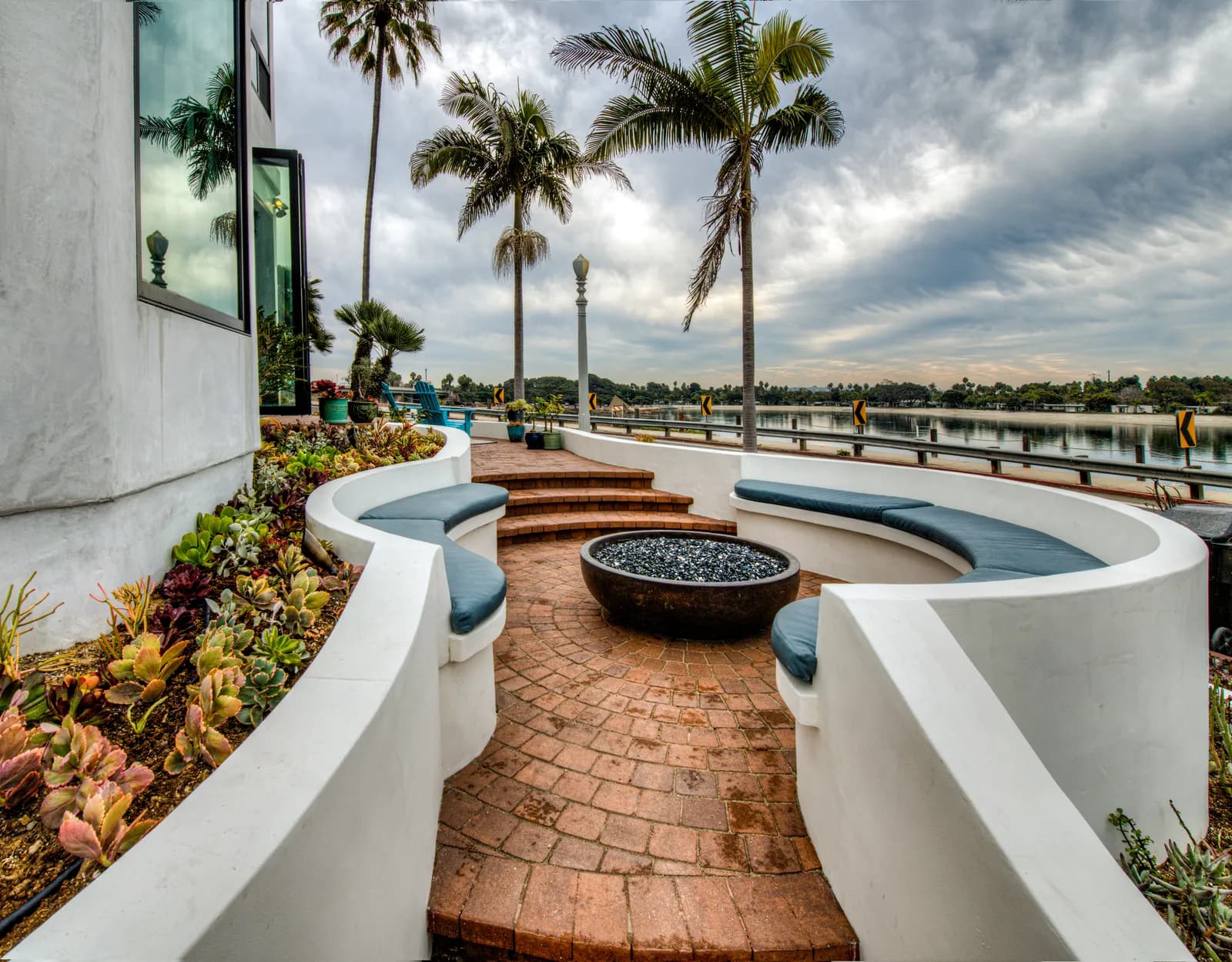 A curved built-in bench with blue cushions around a fire bowl on a brick patio, with palm trees and a waterfront view.