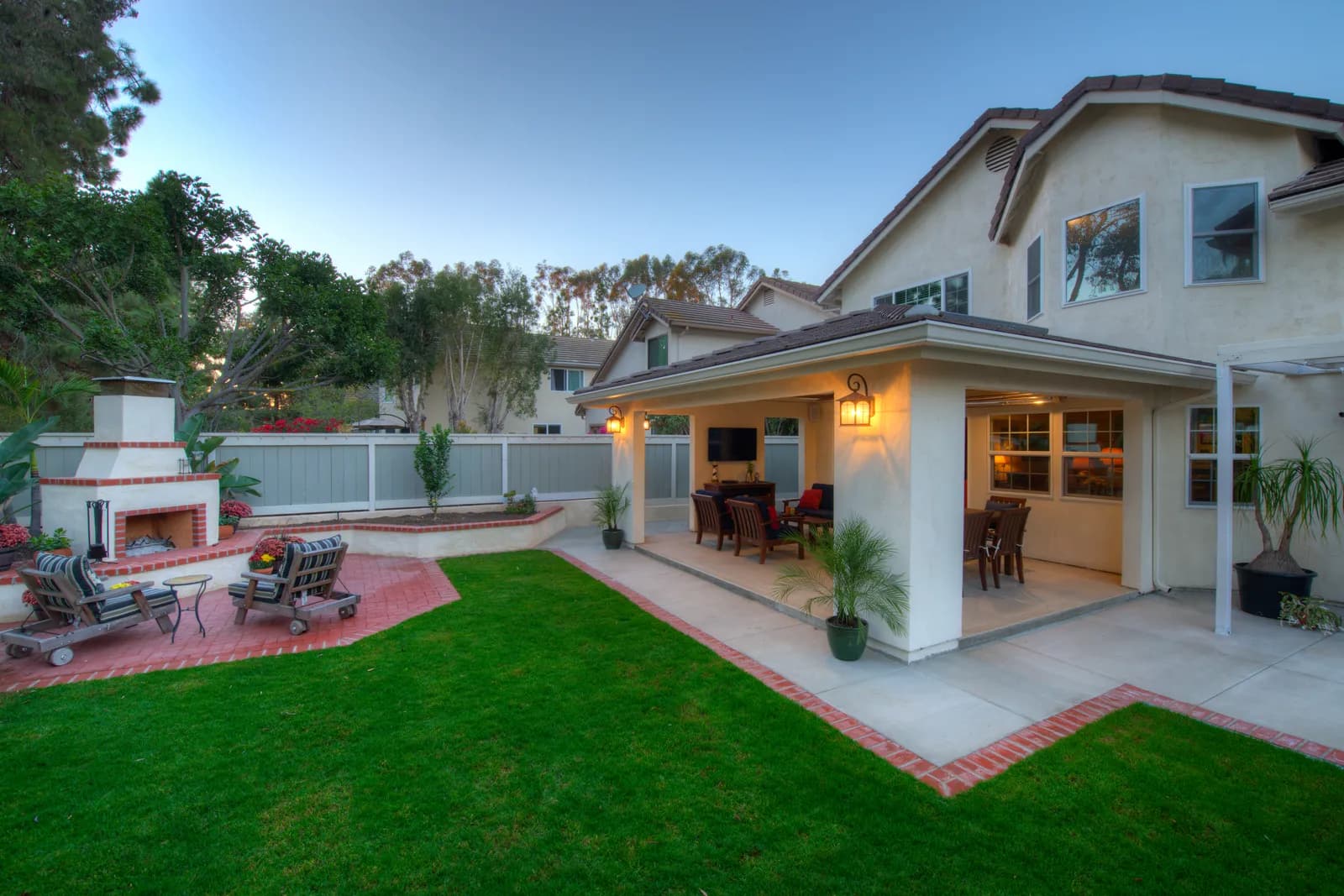 Backyard at dusk with a covered patio addition, outdoor fireplace, brick-edged patio, and manicured lawn.