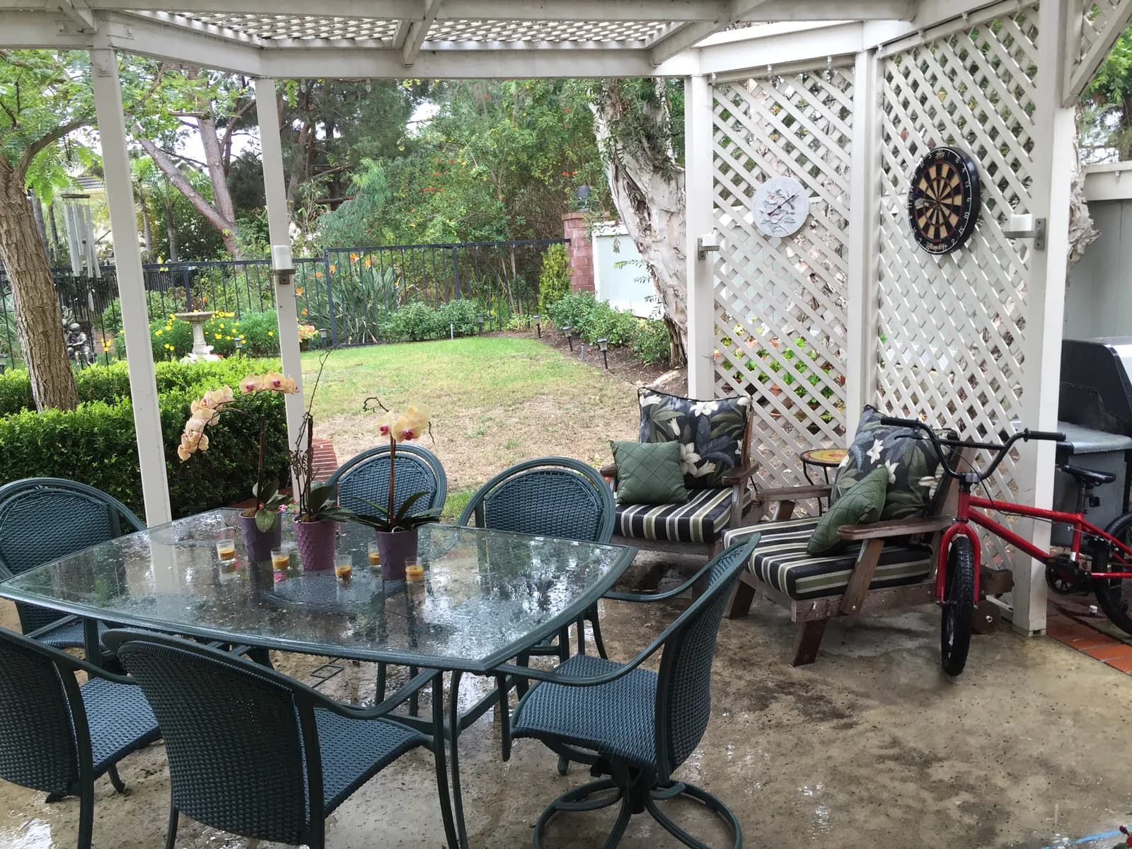 A backyard patio under a white lattice pergola with a glass dining table, wicker chairs, and a lawn beyond.
