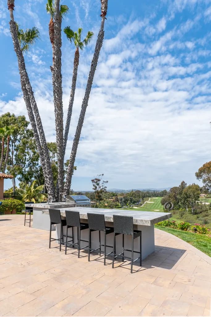 Outdoor concrete bar counter with wicker stools and a grill overlooking a green valley under palm trees.