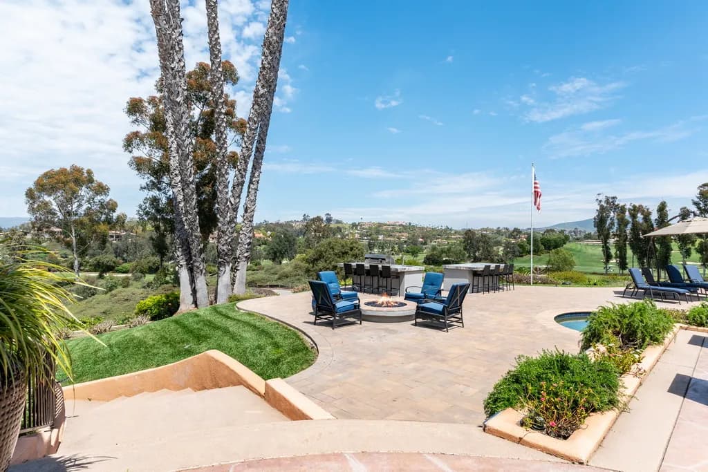 A paver patio with a round fire pit, blue cushioned chairs, an outdoor bar, a pool edge, and a golf course view.