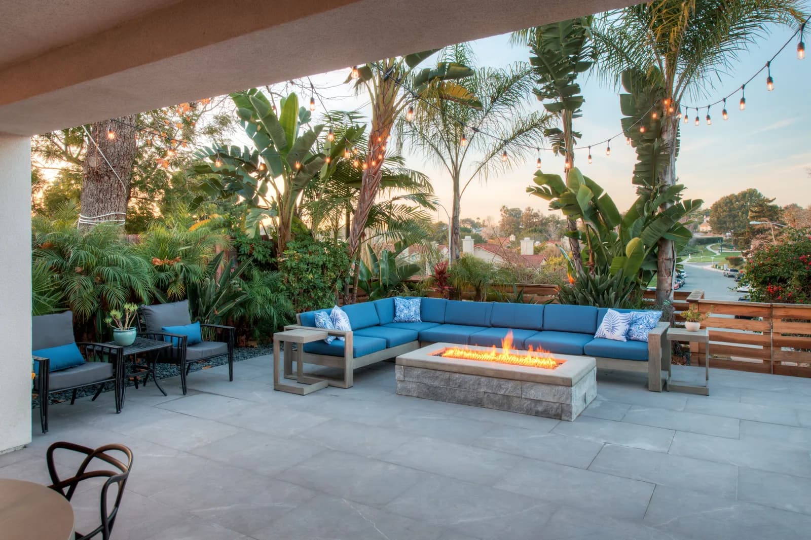 Covered patio at dusk with a blue outdoor sectional around a stone fire pit, string lights, and palm trees.