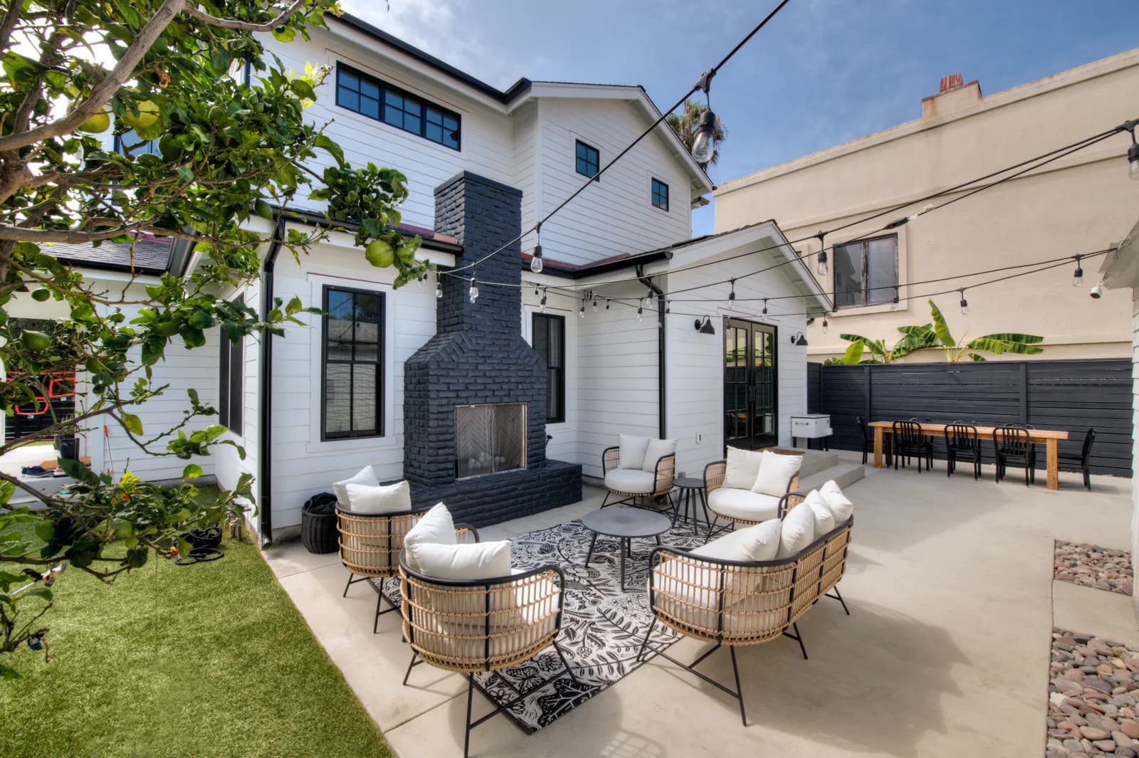 Backyard patio with black brick outdoor fireplace, rattan lounge chairs, string lights, and dining table.