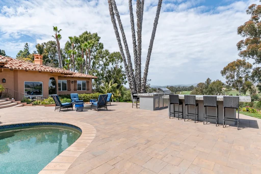 Expansive paver patio with curved pool edge, outdoor kitchen island with barstools, palm trees, and valley views.