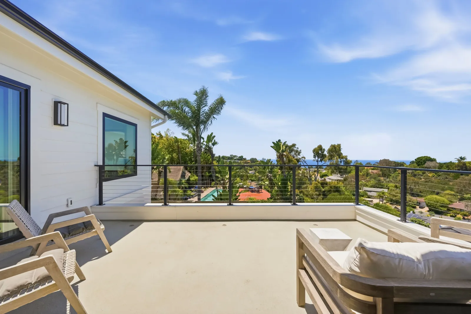 A rooftop deck with cable railing, wood lounge furniture, and a view over palm trees to the ocean.