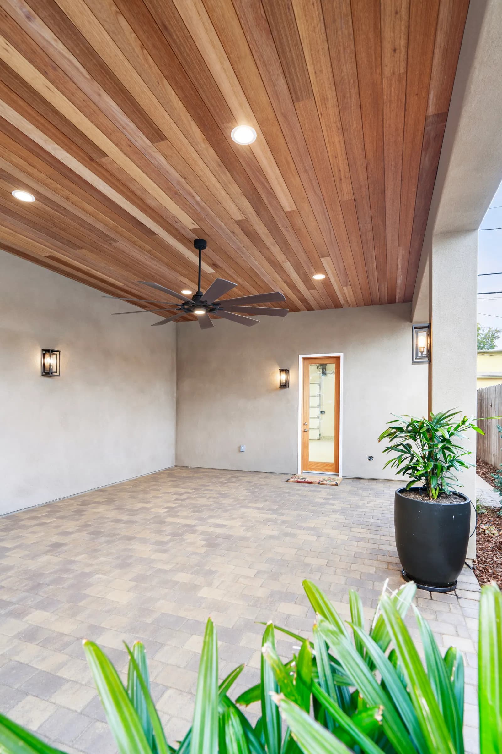 A covered patio with a stained wood plank ceiling, large ceiling fan, paver floor, and potted palm.