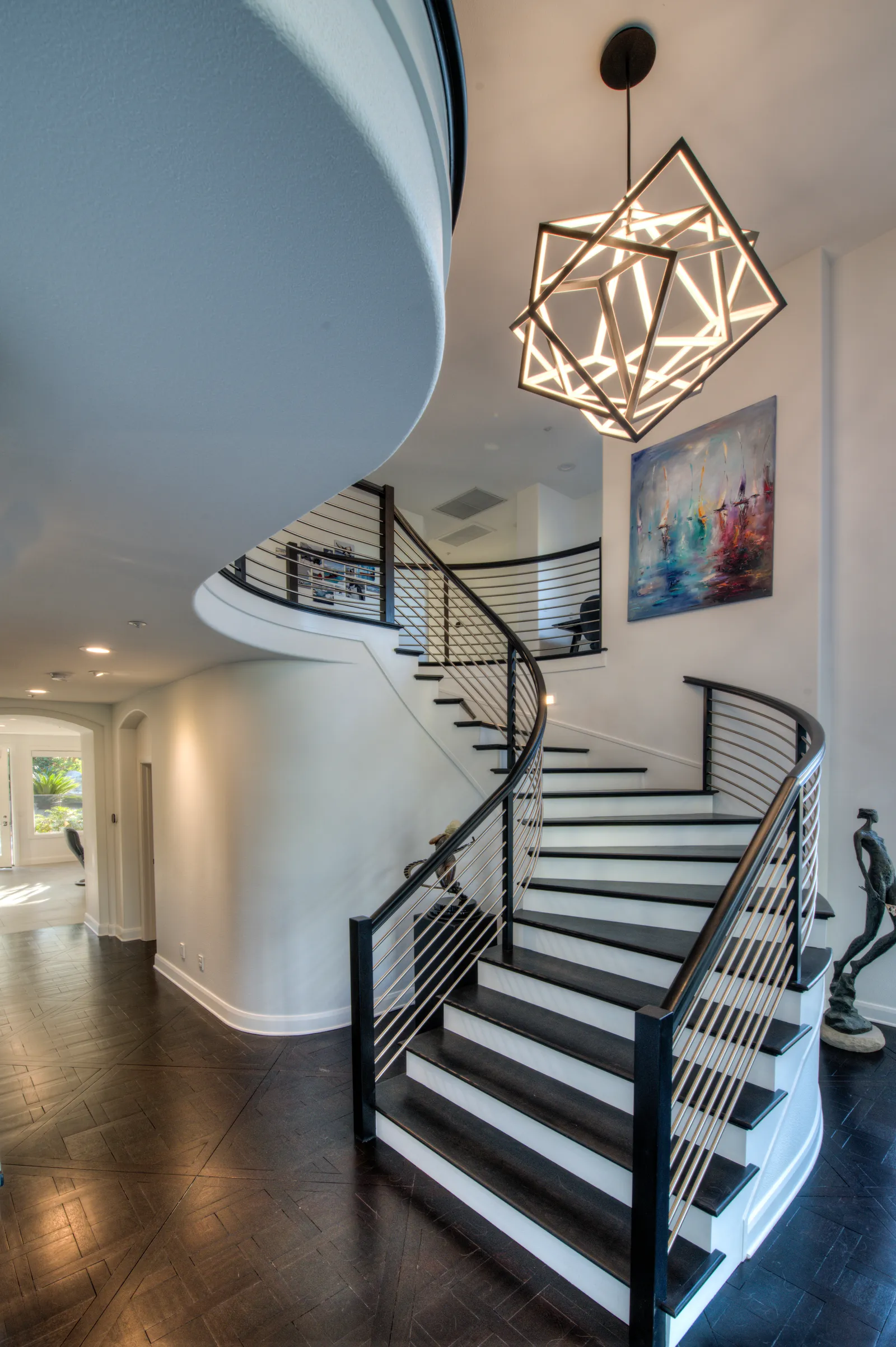 A curved staircase with dark treads, metal rail, and a geometric cube chandelier in a two-story foyer.