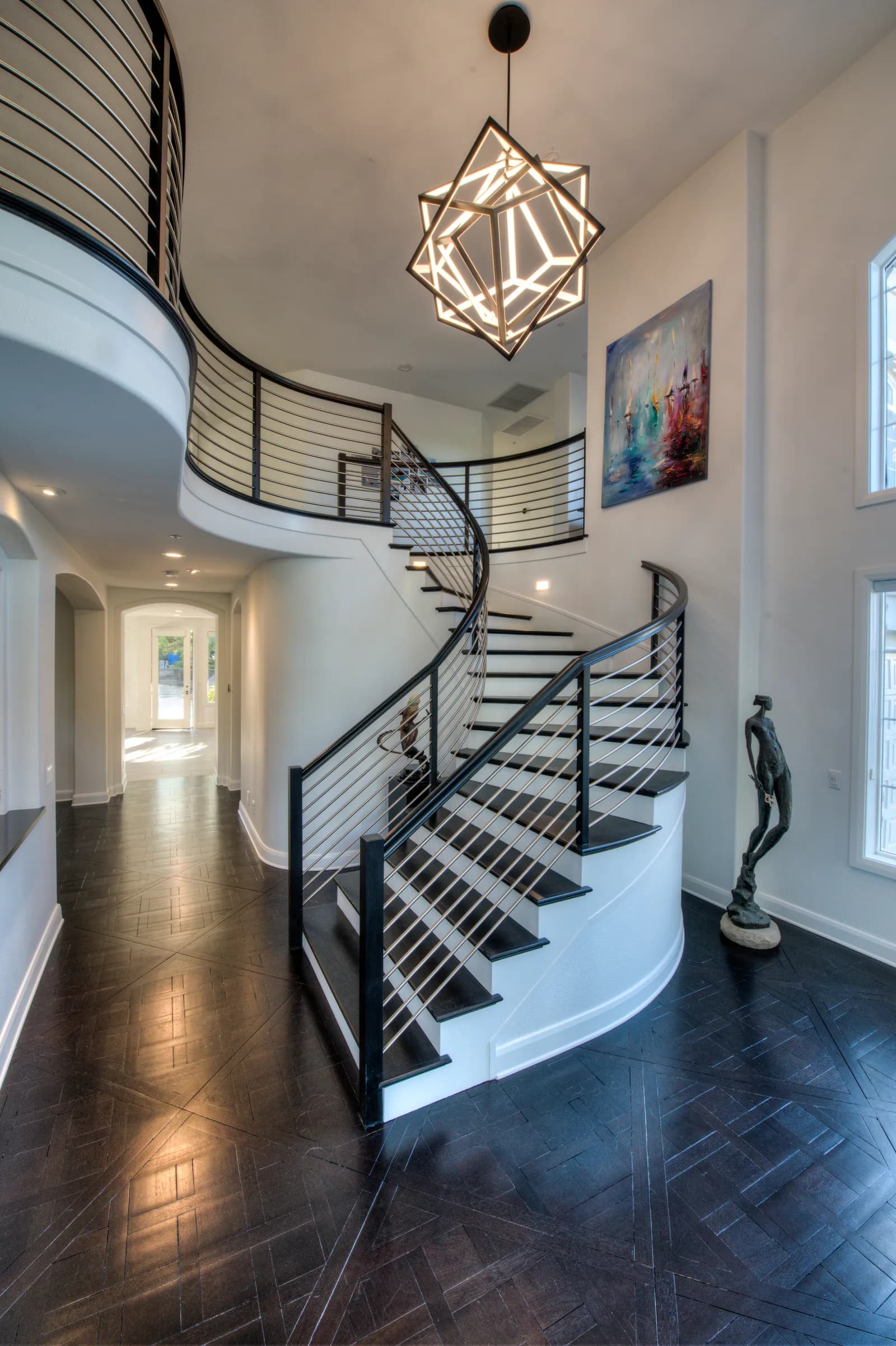 Two-story foyer with a curved staircase, horizontal rail balusters, geometric pendant light, and dark parquet floor.