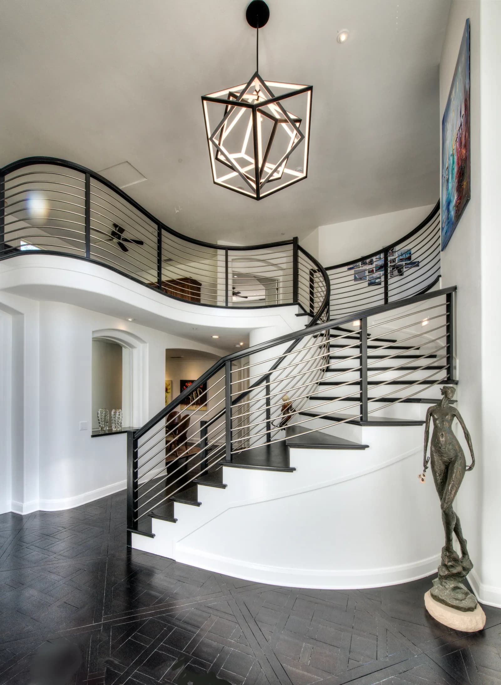 A two-story foyer with a curving staircase, steel rod railings, a geometric chandelier, and a bronze sculpture.