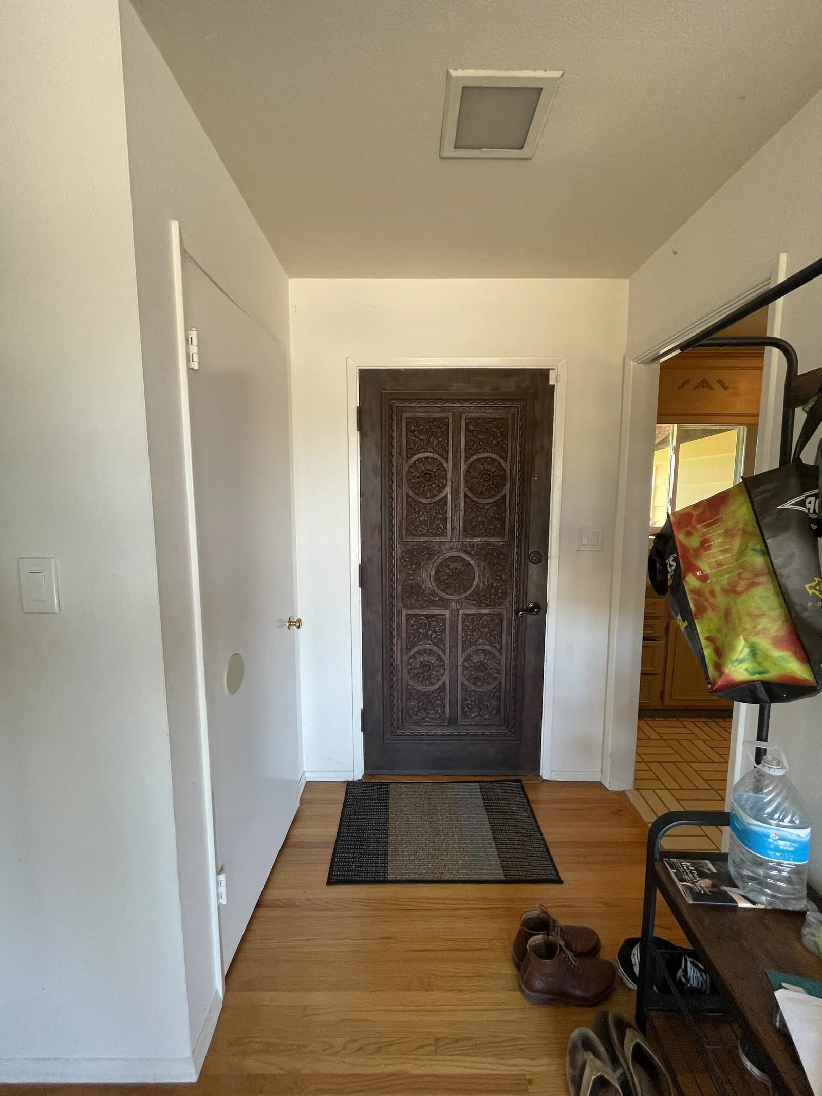 A small entry hall with an ornately carved dark wood front door, oak flooring, an area rug and shoes and bags along the side.
