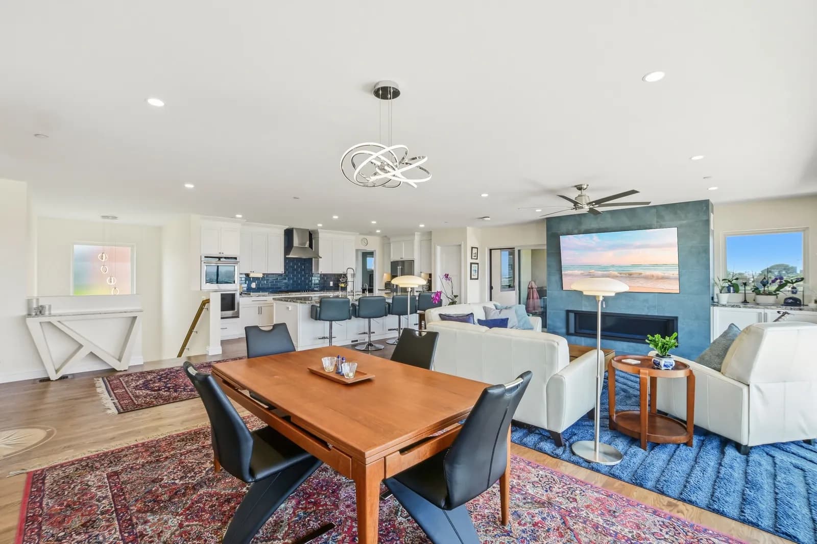 Open-plan interior with dining table in front of a tile fireplace wall, white kitchen with blue subway backsplash, and layered area rugs.