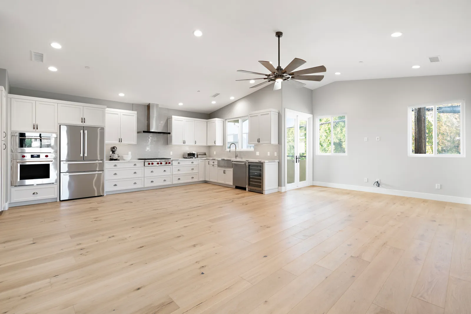 A wide open great room with vaulted ceiling, a large ceiling fan, wide-plank oak floors, and a white kitchen with stainless appliances along the far wall.
