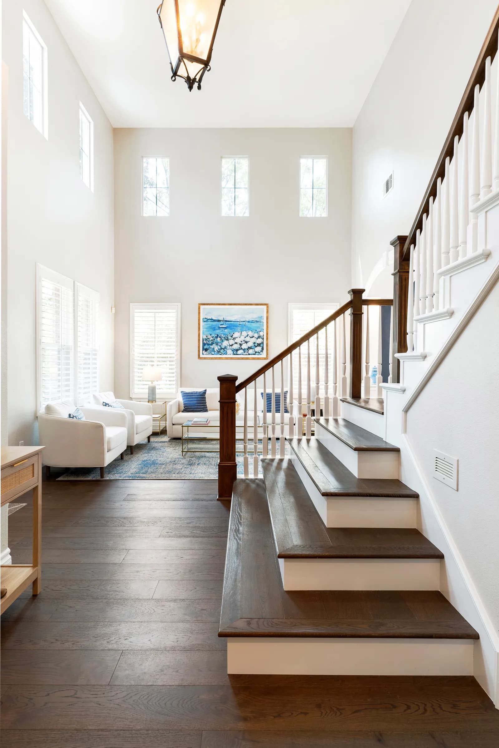 Two-story entry with a dark wood staircase and white balusters overlooking a staged sitting room with tall windows.