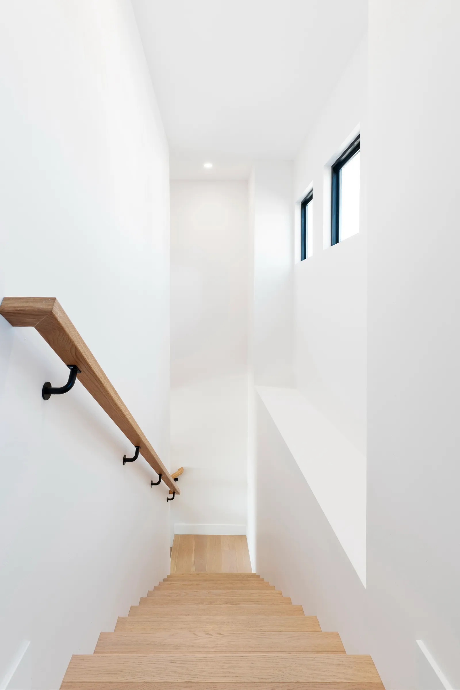 Bright white stairwell with oak treads, a wall-mounted oak handrail on black brackets, and clerestory windows.